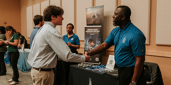 A young man in a dress shirt handshaking a recruiter in a blue polo at a career fair exhibit table.